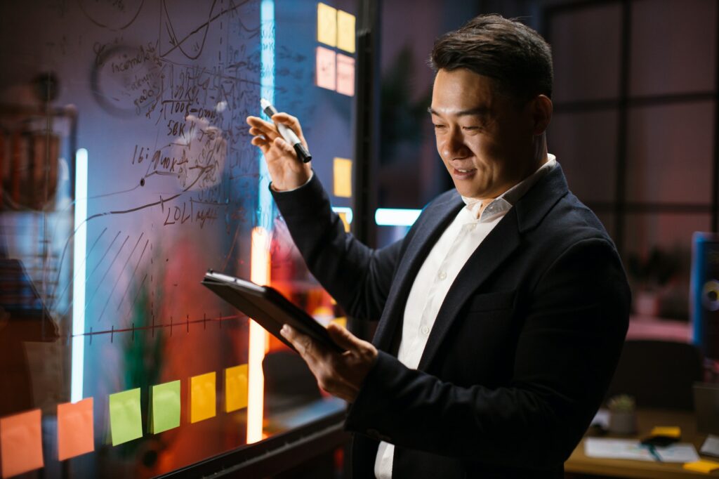 Korean businessman writing on glass board with attached sticky notes while looking at digital tablet